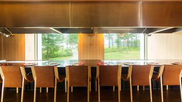 A modern dining area with a long table, six chairs, and a view of a green landscape through large windows.