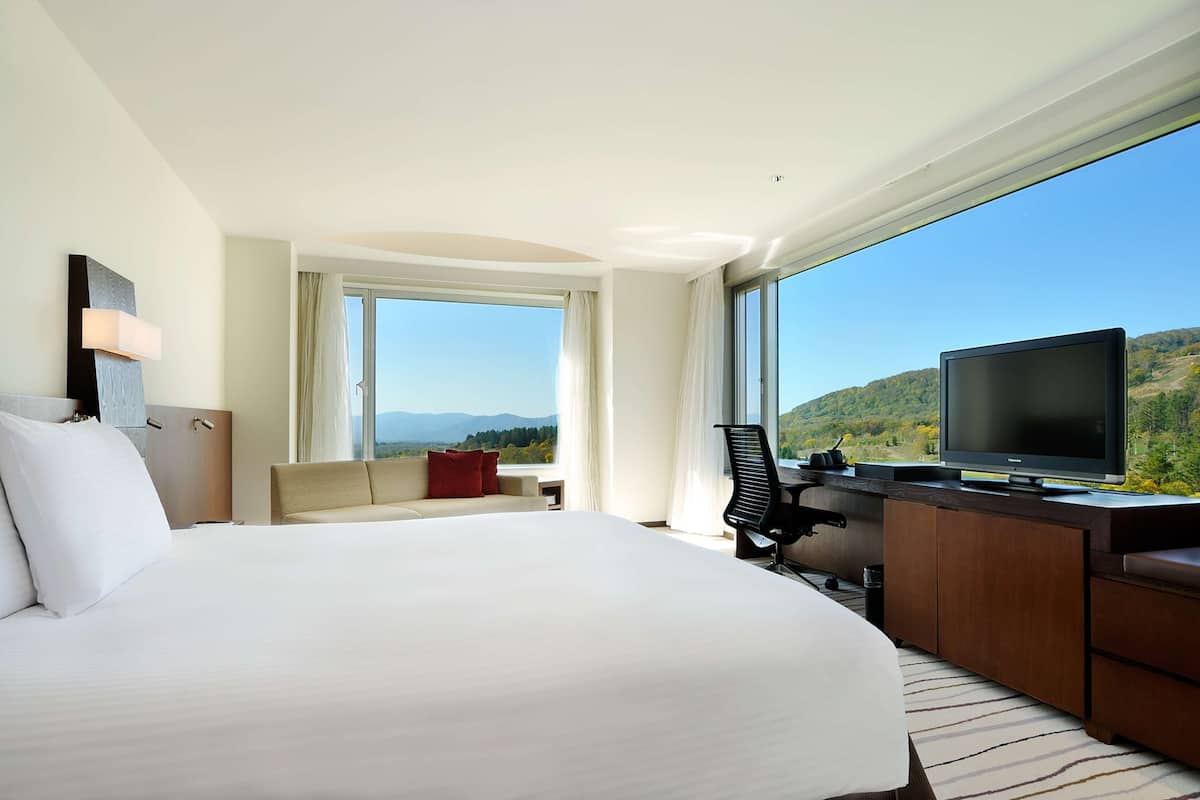 Ein Hotelzimmer mit einem großen Bett, einem Schreibtisch mit Fernseher, einem Sessel und einer Couch. Ein Fenster bietet Blick auf Berge und Grünfläche.