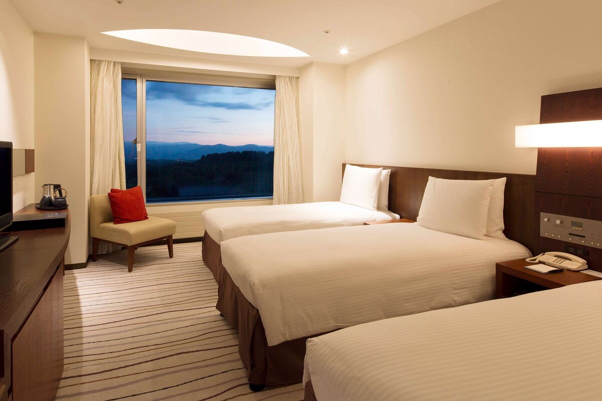 A hotel room with two beds, a TV, a coffee maker, and a view of mountains through the window.