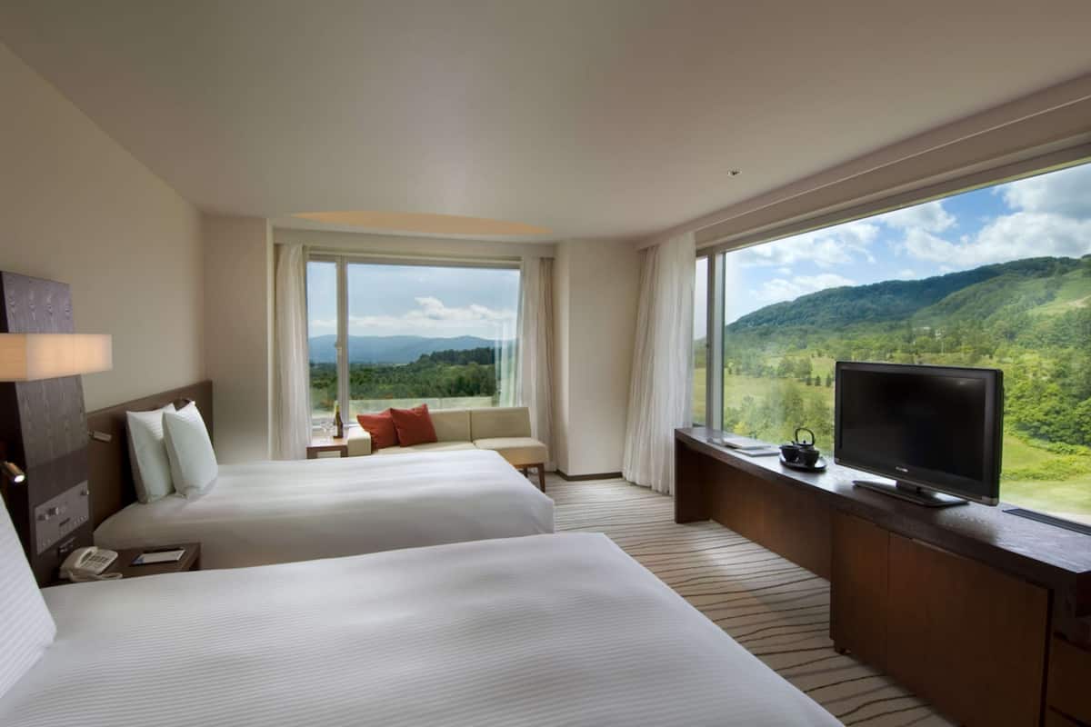 Ein Hotelzimmer mit zwei Betten, einem Fernseher, einem Schreibtisch und Blick auf eine gebirgige Landschaft.