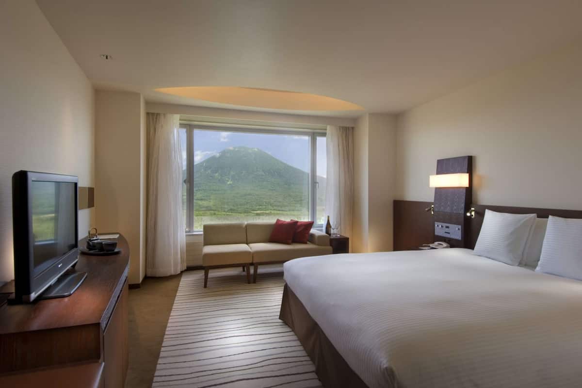 Ein Hotelzimmer mit einem großen Bett, einem Fernseher auf einer Holzkombie, einer Couch, einem Fenster mit Bergblick und einem Nachttisch mit Lampe.