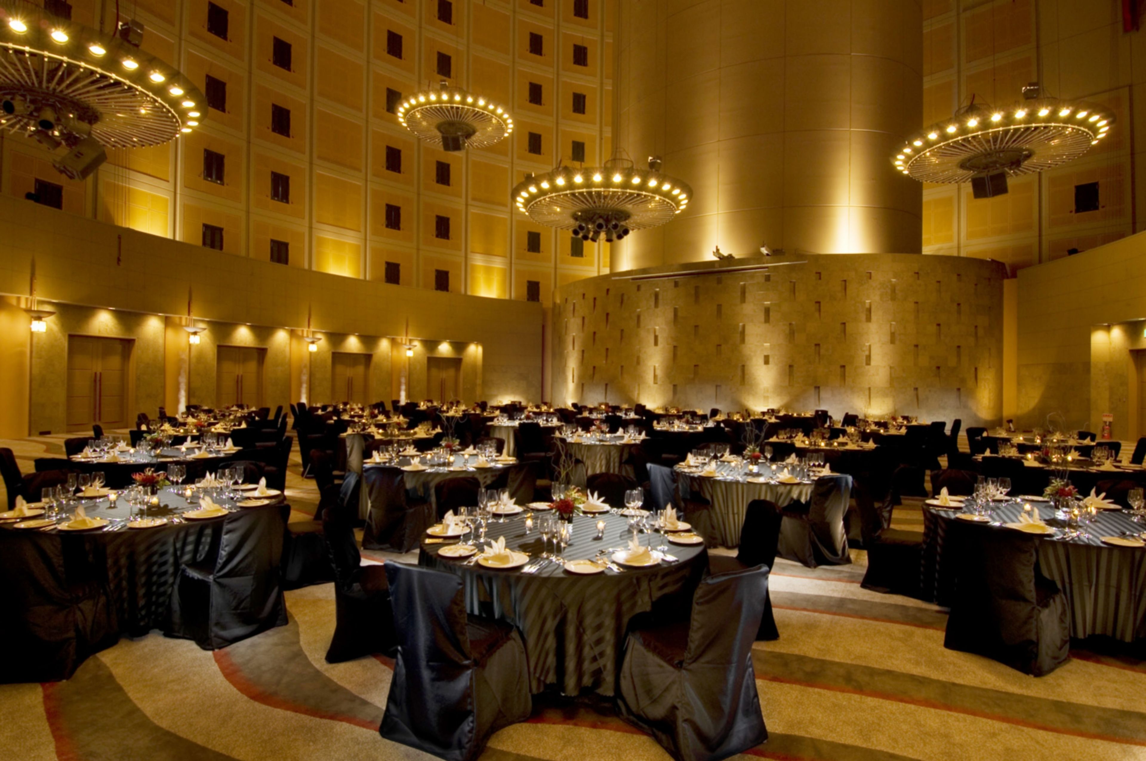 Une vaste salle de banquet, dotée de nombreuses tables rondes dressées pour le repas, de hauts plafonds et d’appareils d’éclairage décoratifs.