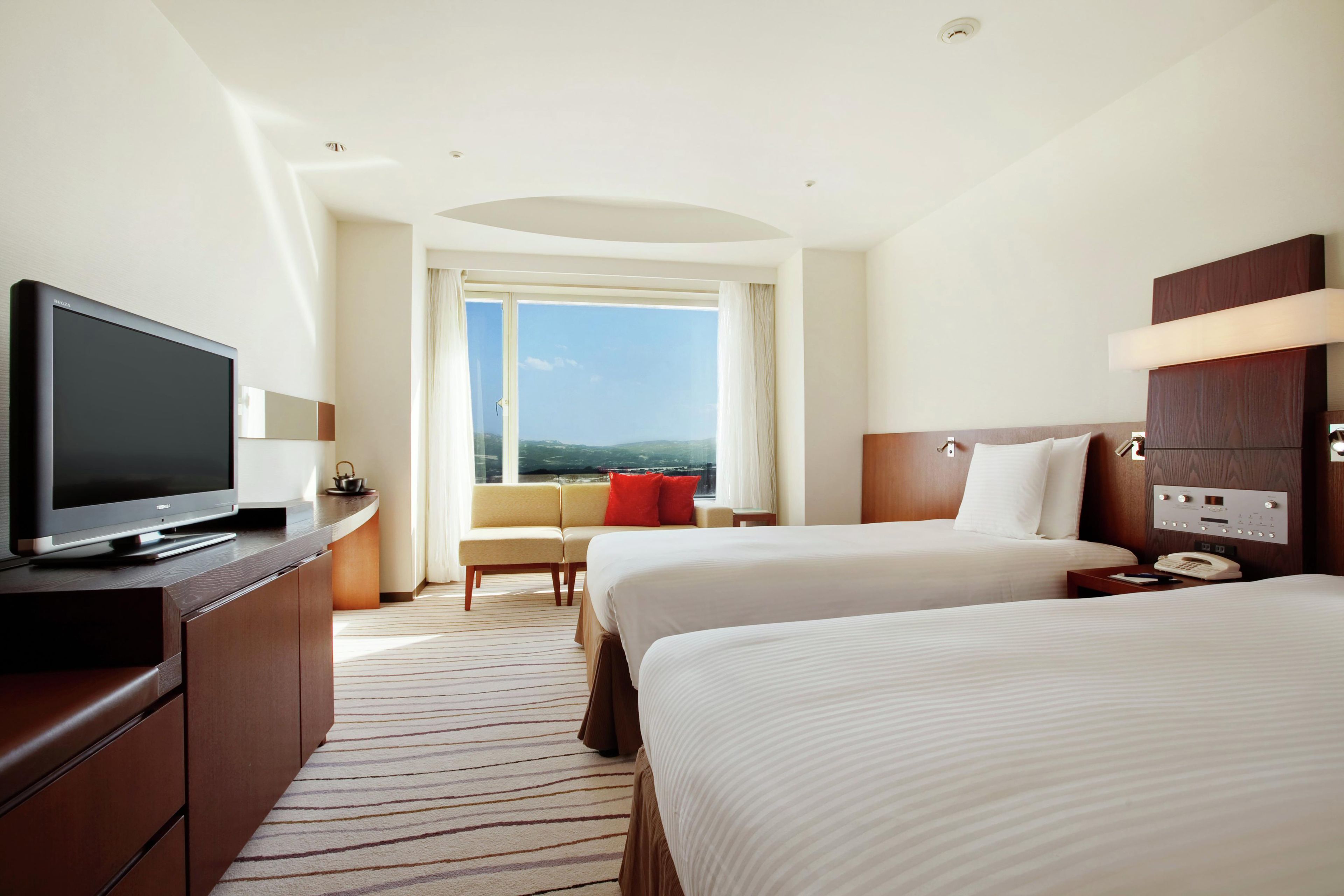 Quarto de hotel com televisão, sofá, duas camas, janela com vista e painel de controle.
