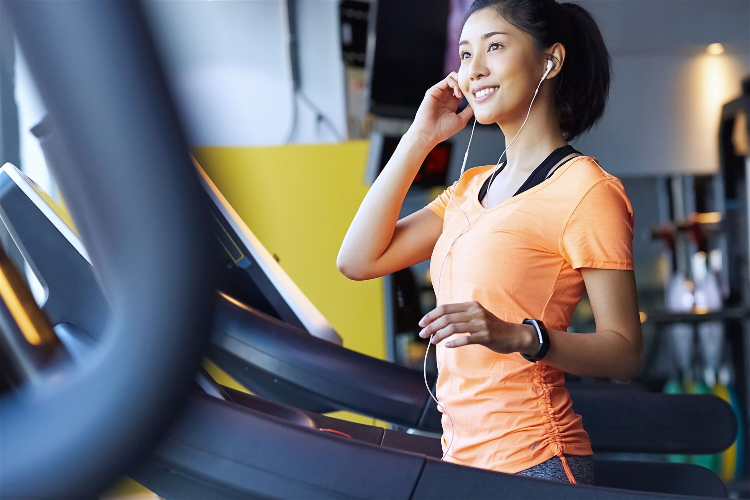 Eine Person läuft auf einem Laufband in einem Fitnessstudio mit Trainingsgeräten. Im Hintergrund ist eine gelbe Wand zu sehen.