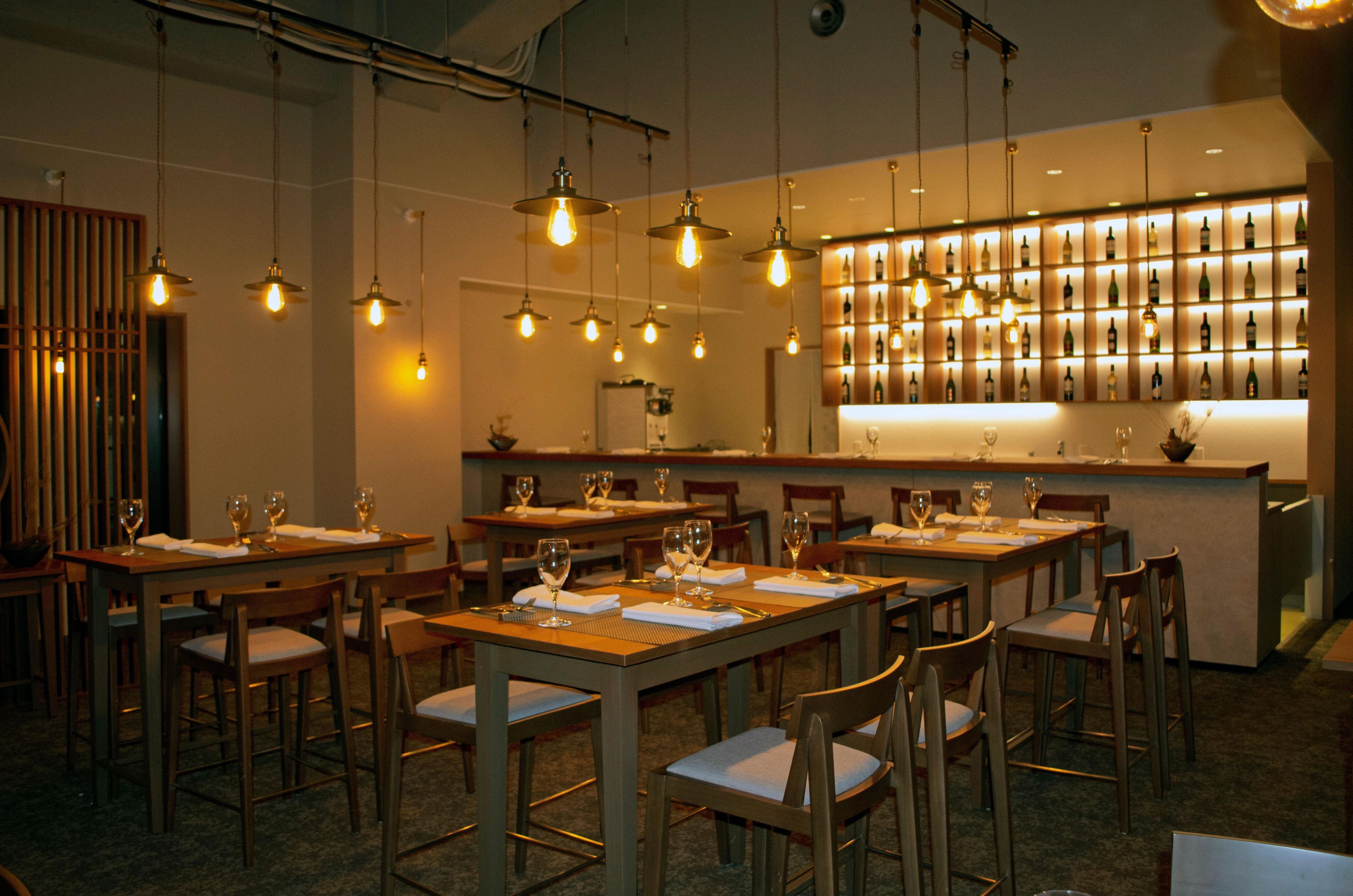 Ein Restaurantinneres mit mehreren Tischen, Stühlen, hängenden Pendelleuchten und einer Weinwand.