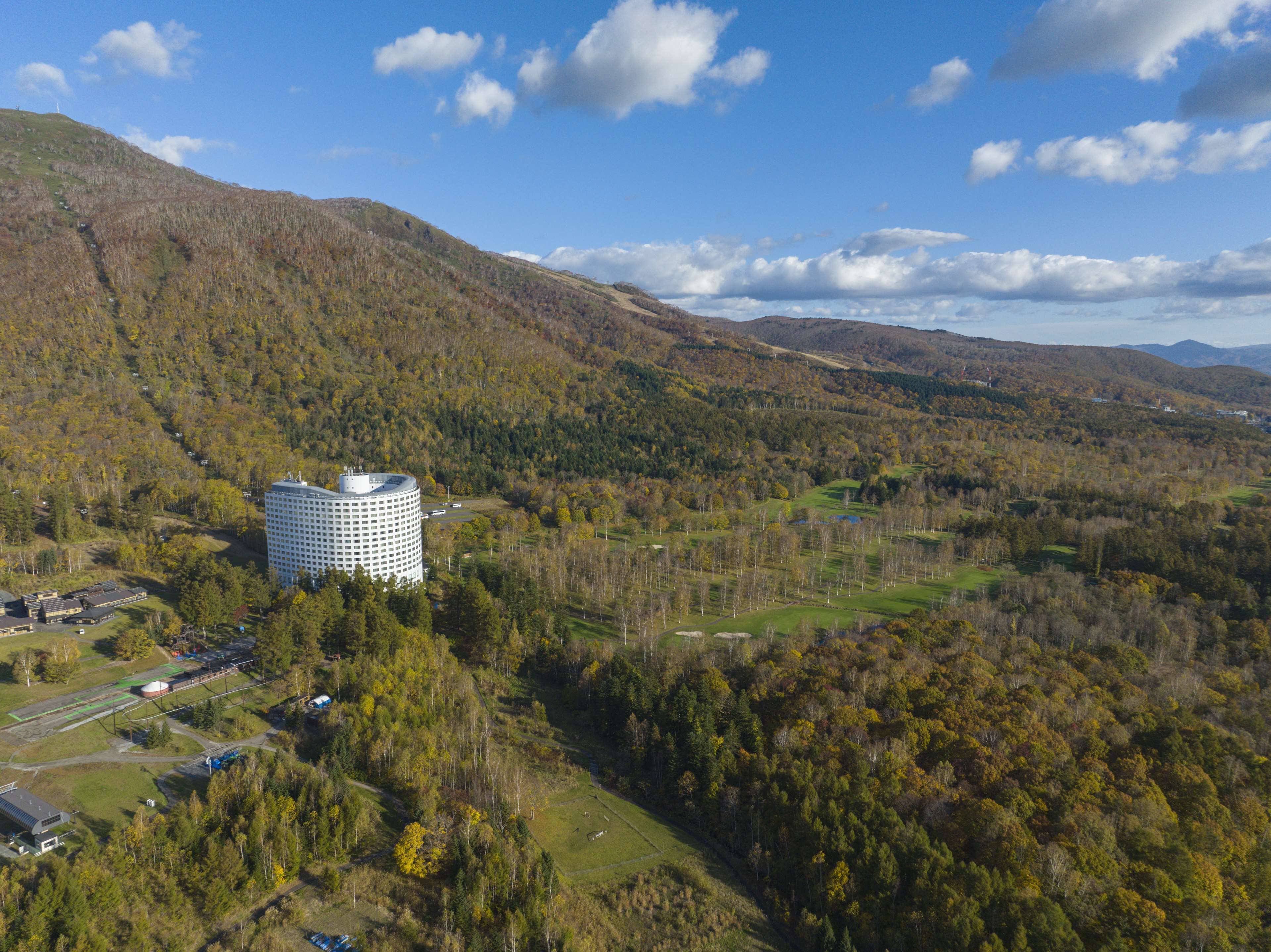 Un hotel grande de varios pisos con diseño circular, rodeado de un paisaje boscoso y colinas.