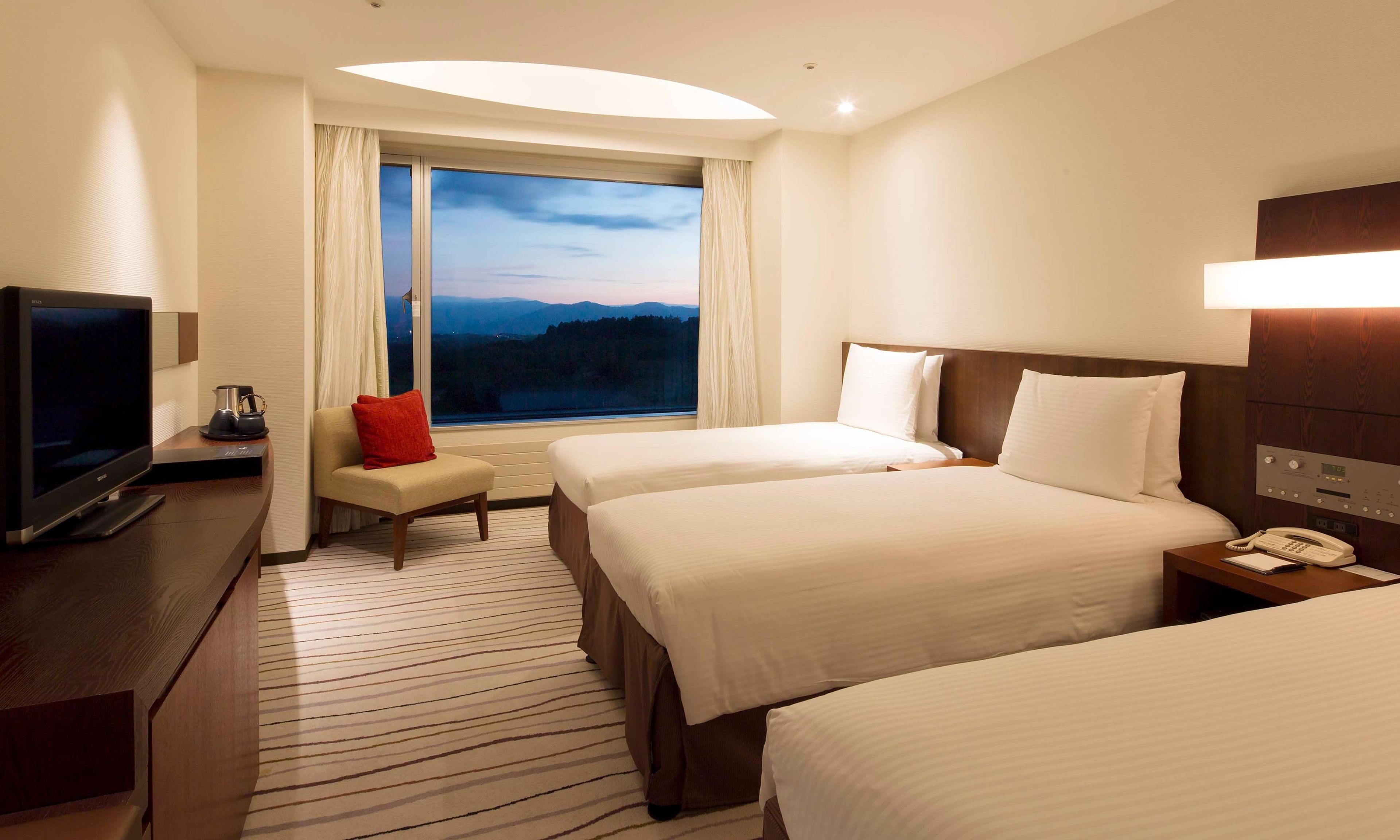 Habitación de hotel con dos camas, un televisor, una mesita de noche con teléfono y una ventana con vistas a la montaña.