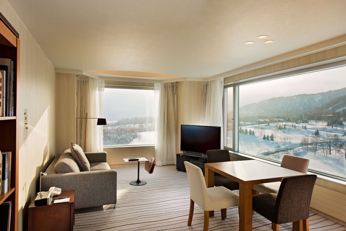 Ein modernes Hotelzimmer mit großem Fenster, einer Couch, einem Esstisch und einem Flachbildfernseher.
