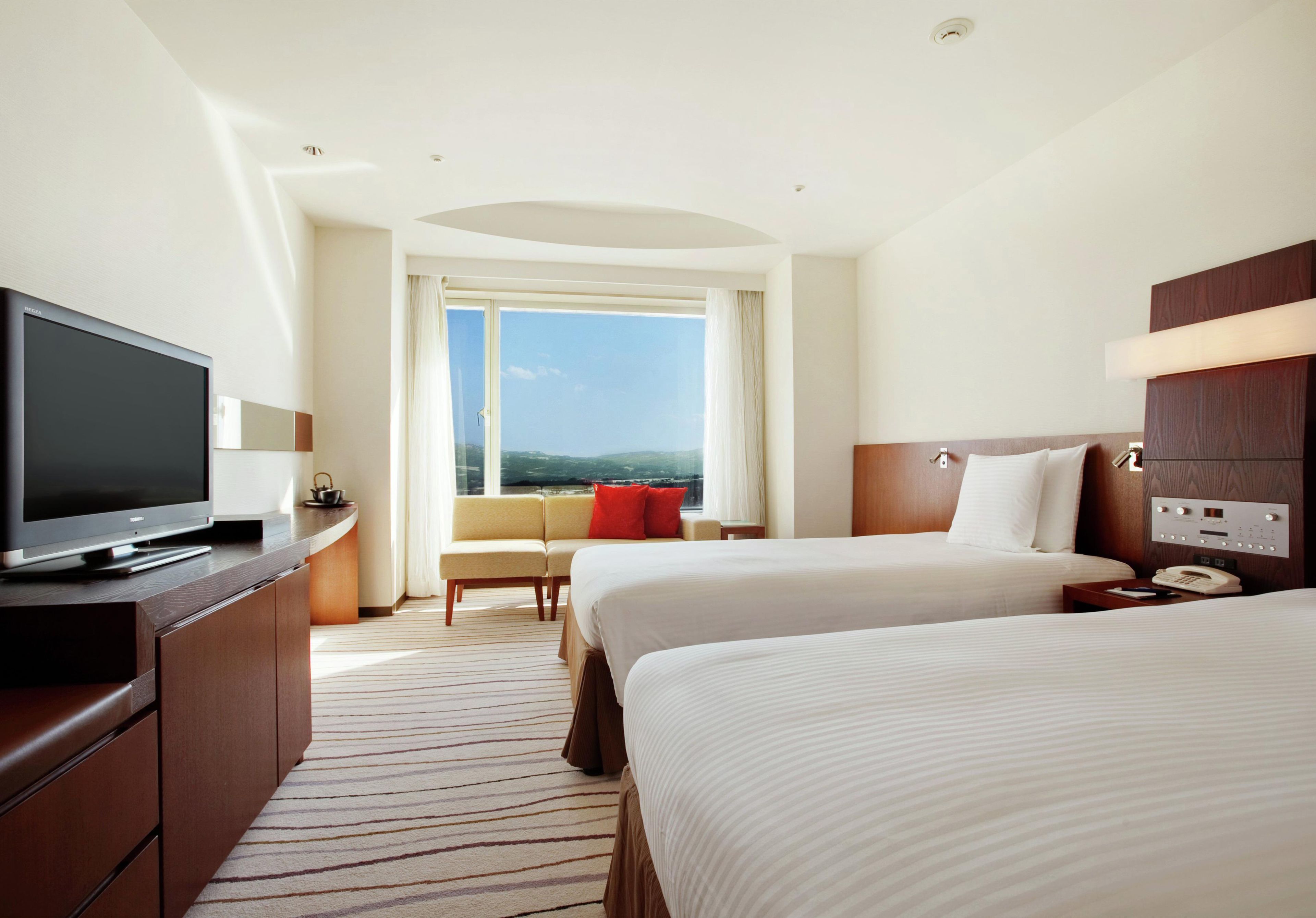 A hotel room with a television, a sofa, two beds, a window with a view, and a control panel.