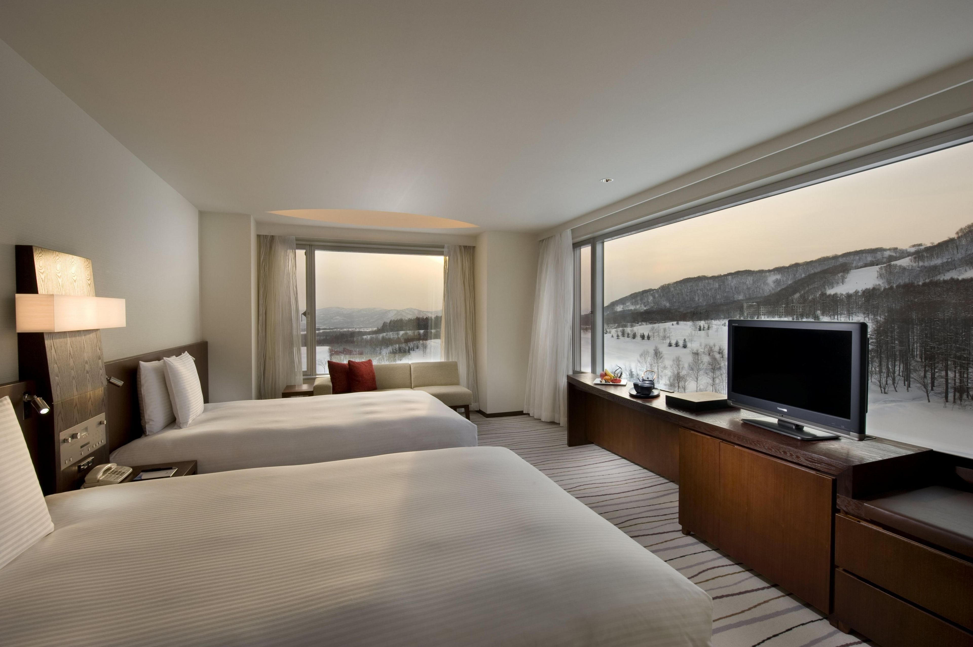 A hotel room with two beds, a TV, a desk, and a large window with a view of a snowy landscape.