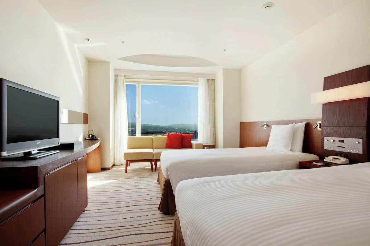 Ein Hotelzimmer mit zwei Betten, einem Fernseher, einer Couch, einem großen Fenster mit Aussicht und einem Holz-Kopfteil.