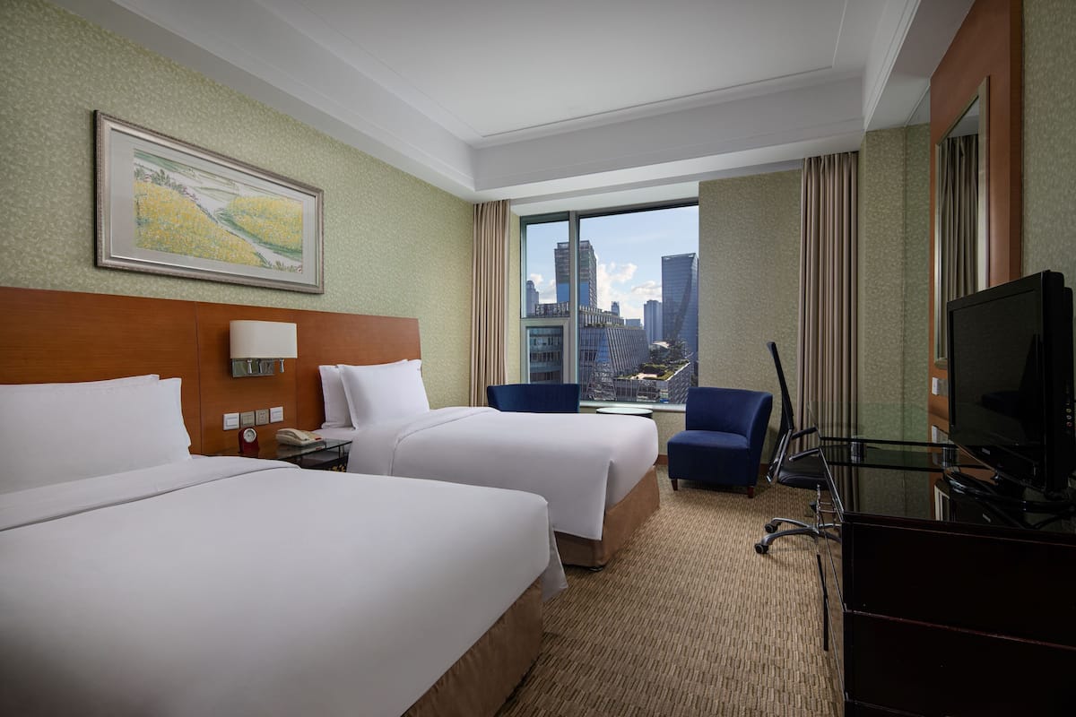 A hotel room with two beds, a desk, a chair, a television, and a large window offering a city view.