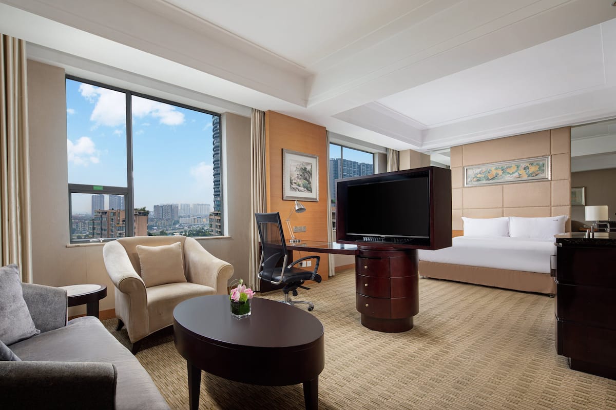 A hotel room with a large bed, a sofa, a desk, a TV, and a view of the city.