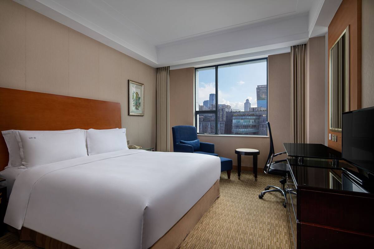 A hotel room with a large bed, a desk with a chair, a television, and a window with a city view.