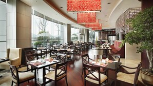 Un restaurant spacieux avec de hauts plafonds, de grandes fenêtres et un décor moderne. Les tables sont disposées avec soin et accompagnées de chaises, et des panneaux décoratifs rouges sont suspendus au plafond.