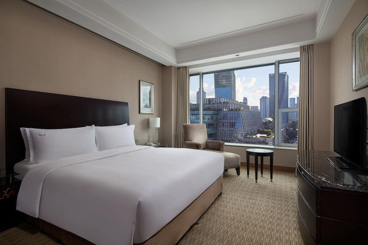 A hotel room with a large bed, a TV, a chair, a small table, and a view of the cityscape through large windows.