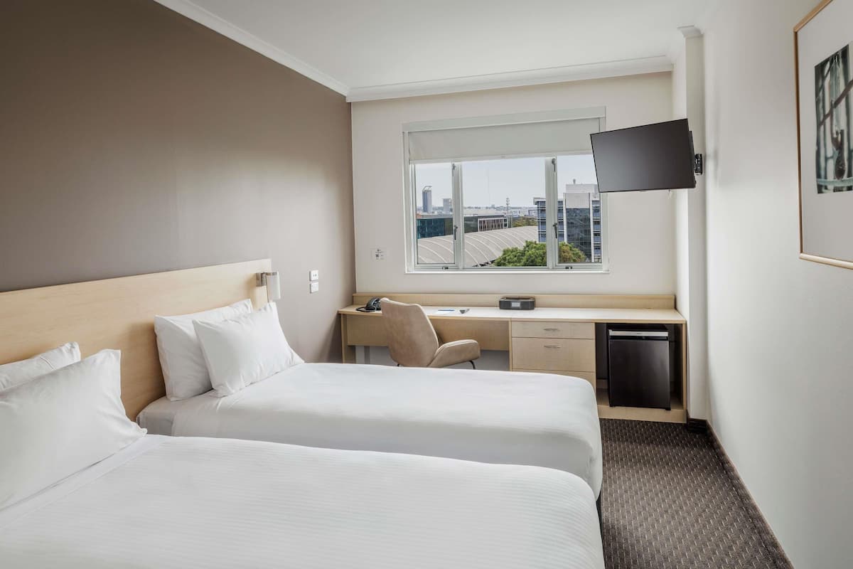 A hotel room with two beds, a desk, a chair, a TV, and a window with a city view.