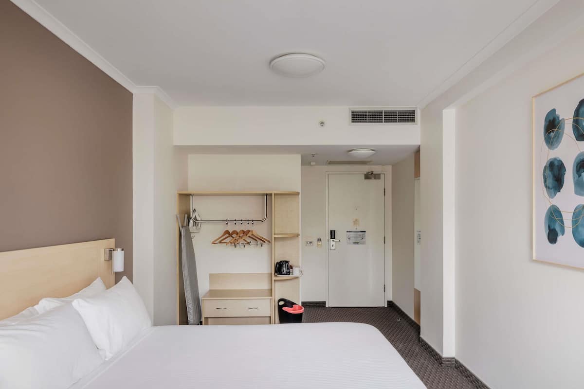 A hotel room with a bed, a wardrobe, a coat rack, a door, and a framed artwork on the wall.