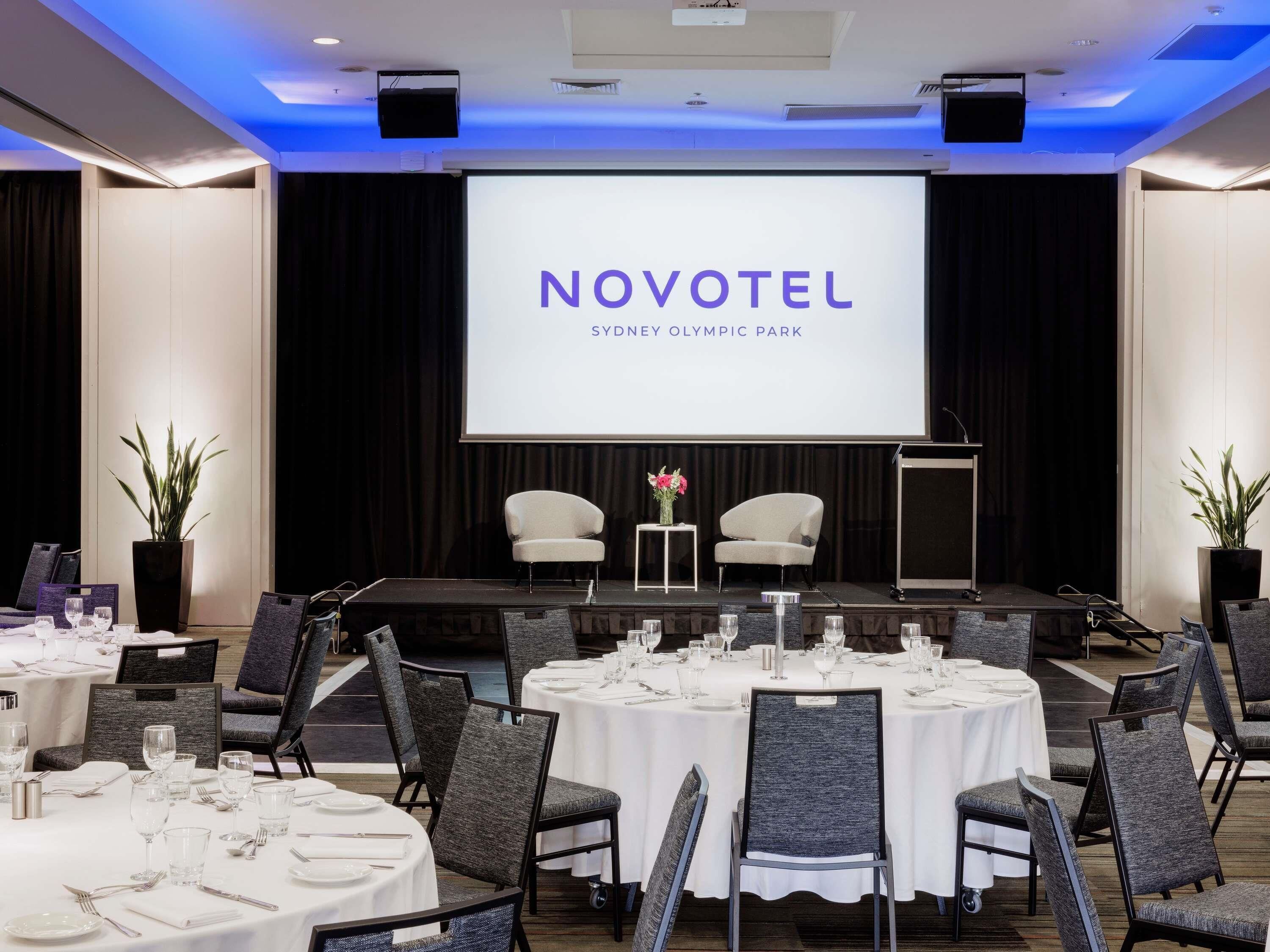 A conference room with a stage, tables set for dining, and a large screen displaying 'NOVOTEL SYDNEY OLYMPIC PARK'.