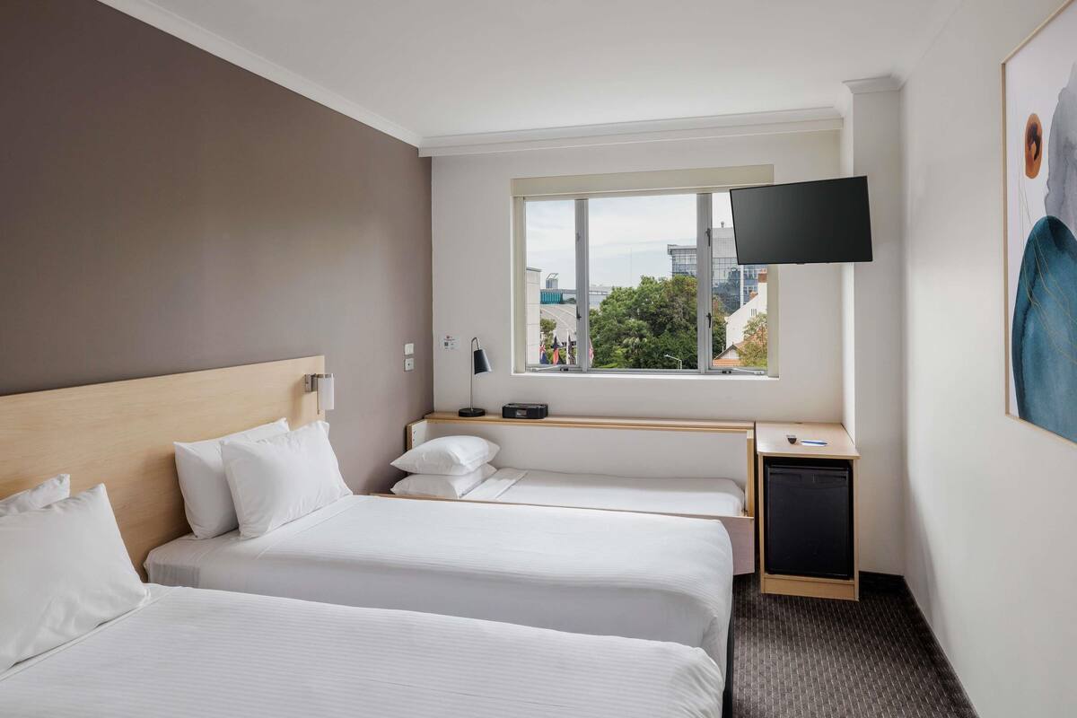 A hotel room with two beds, a desk with a lamp, a TV, and a window with a city view.