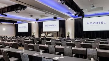 A conference room with rows of tables and chairs, a stage with a screen displaying 'NOVOTEL SYDNEY OLYMPIC PARK', and overhead lighting.
