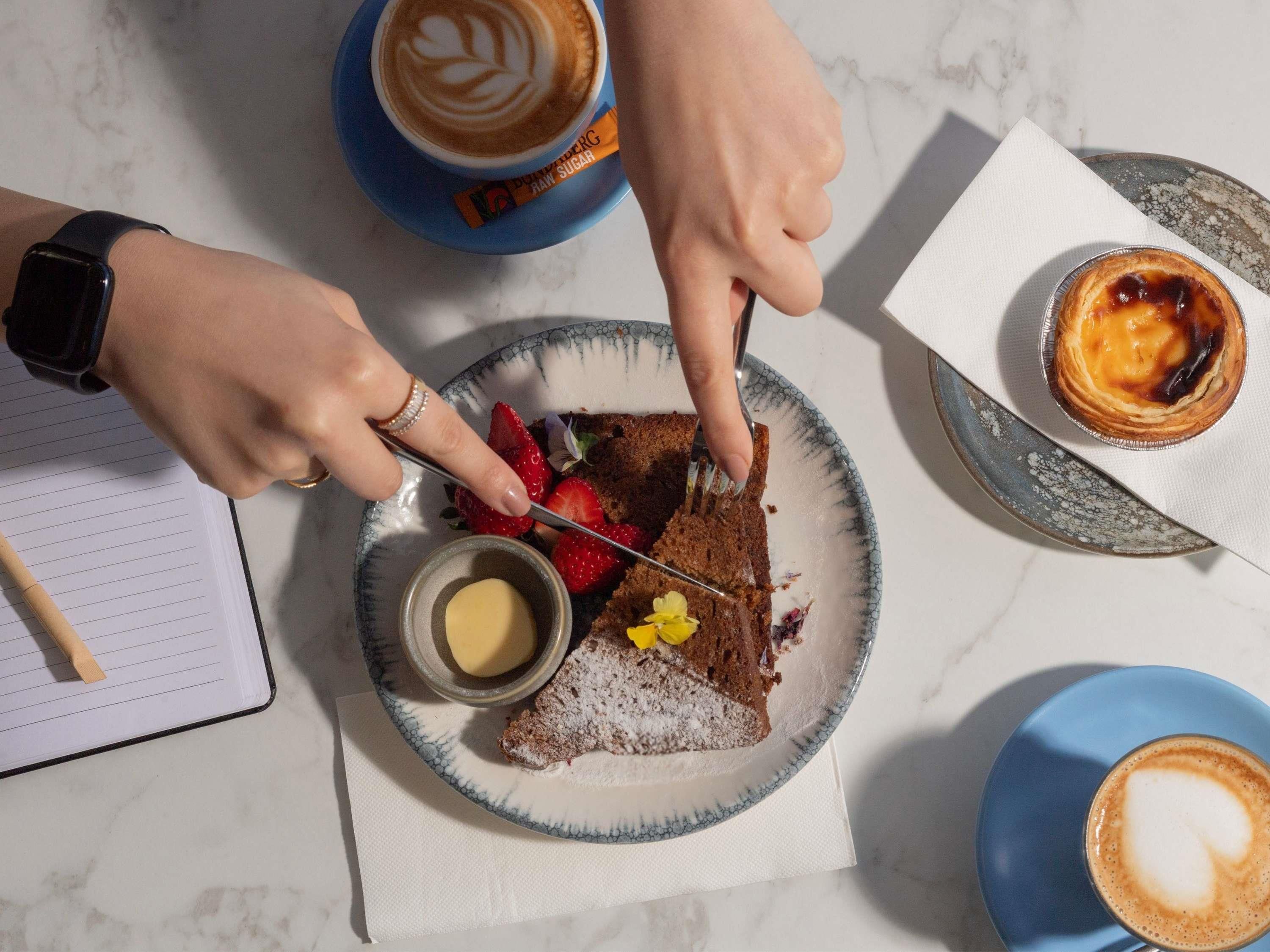 Una persona cortando una rebanada de pastel con fresas y un tazón pequeño de mantequilla en un plato, con una taza de café y un pastelito al fondo.