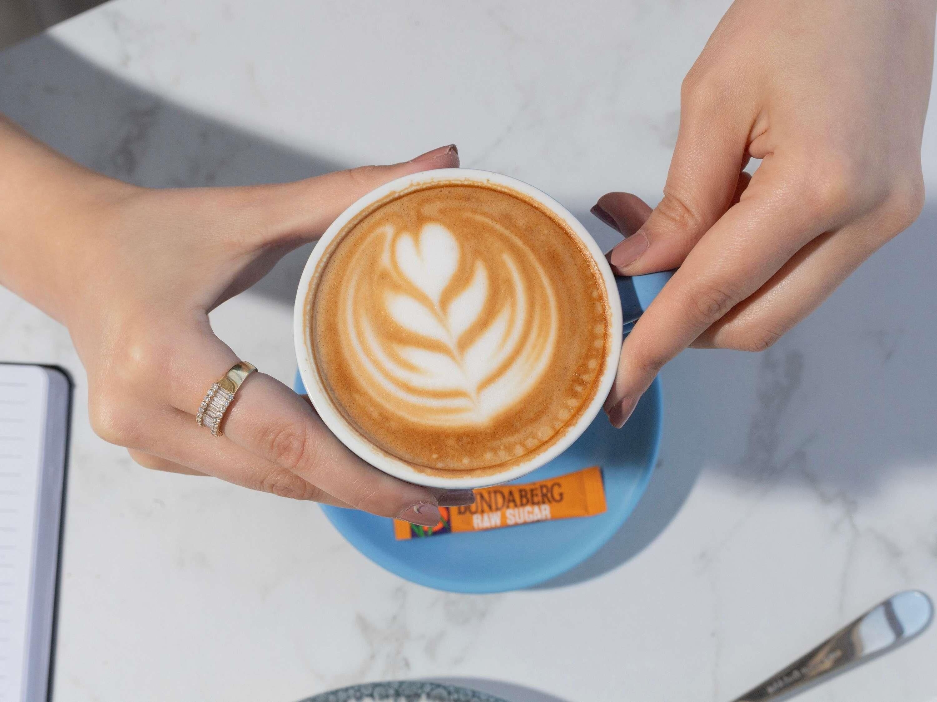A person holding a cup of coffee with a leaf design on the foam, a spoon, and a packet of raw sugar on a blue saucer.