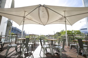 Un área de comedor al aire libre con sillas y mesas de metal, cubierta por un toldo grande.