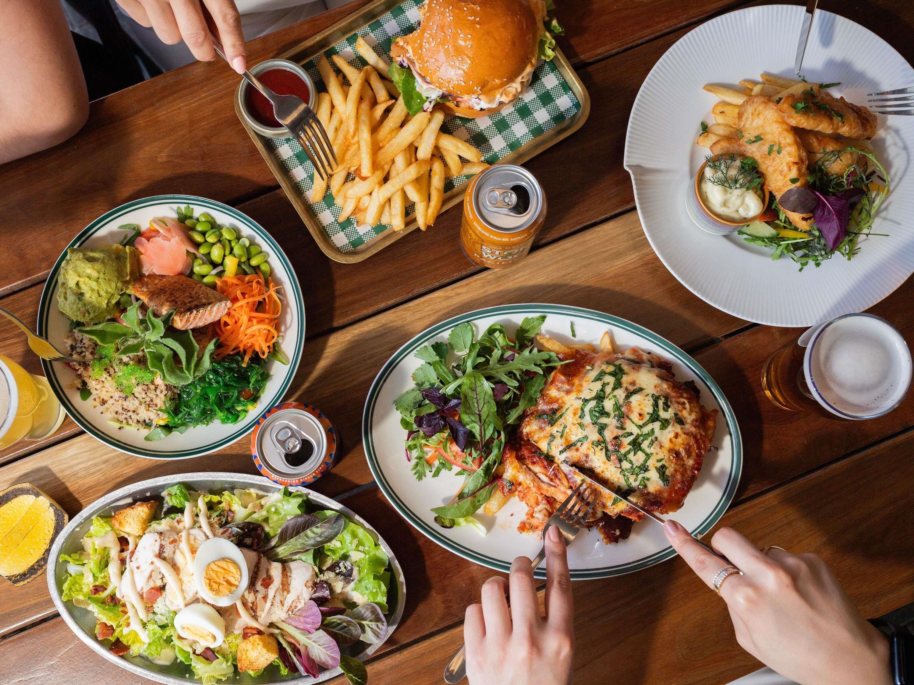 Una mesa con varios platos, incluyendo una hamburguesa, papas fritas, ensalada y un plato de pescado con patatas fritas.