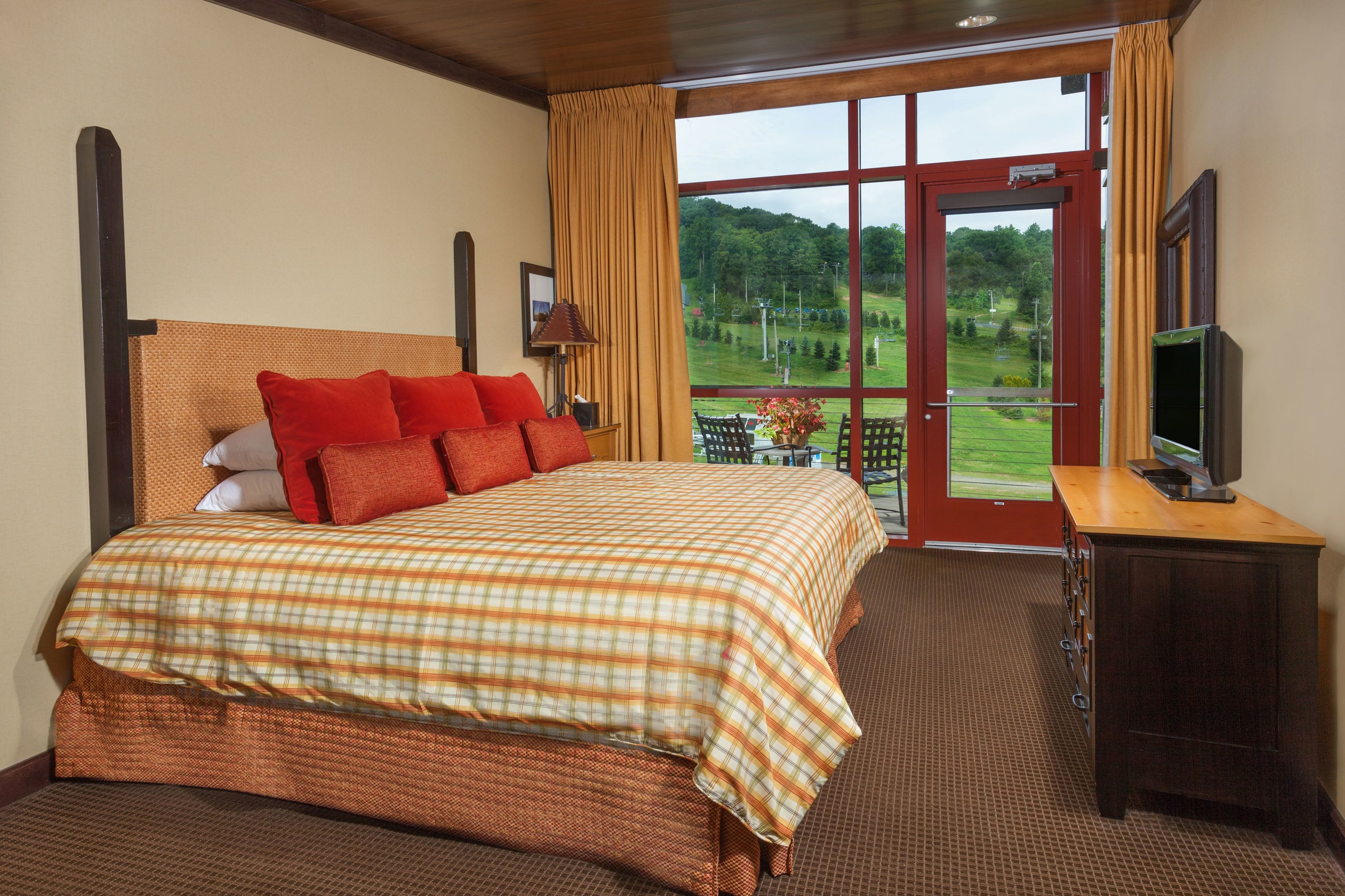 Une chambre d’hôtel avec un grand lit, une commode en bois, une télévision et une vue sur un paysage verdoyant, visible à travers une porte vitrée.