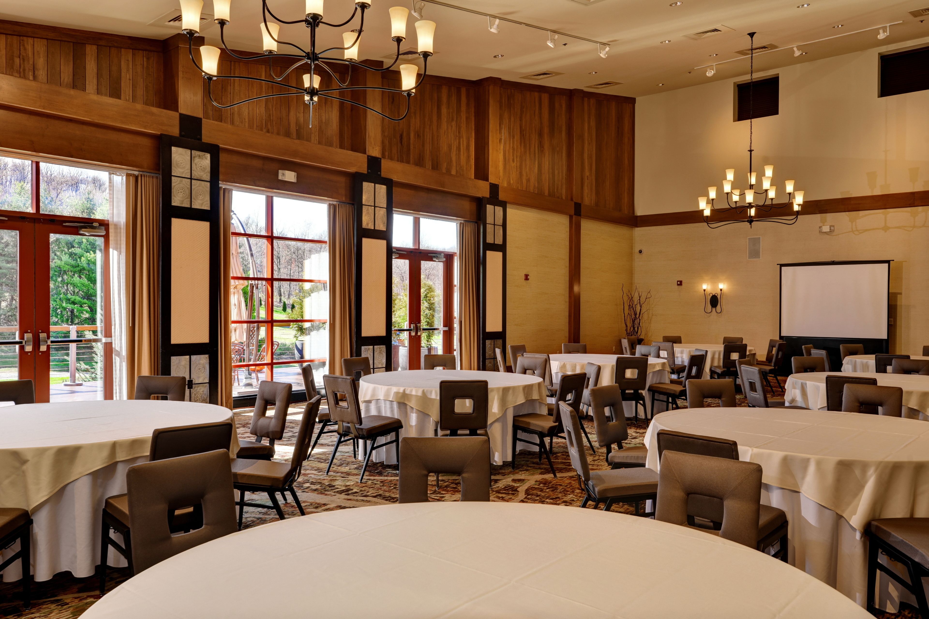 A conference room with multiple round tables, chairs, a projector screen, and a large window with a view of trees.