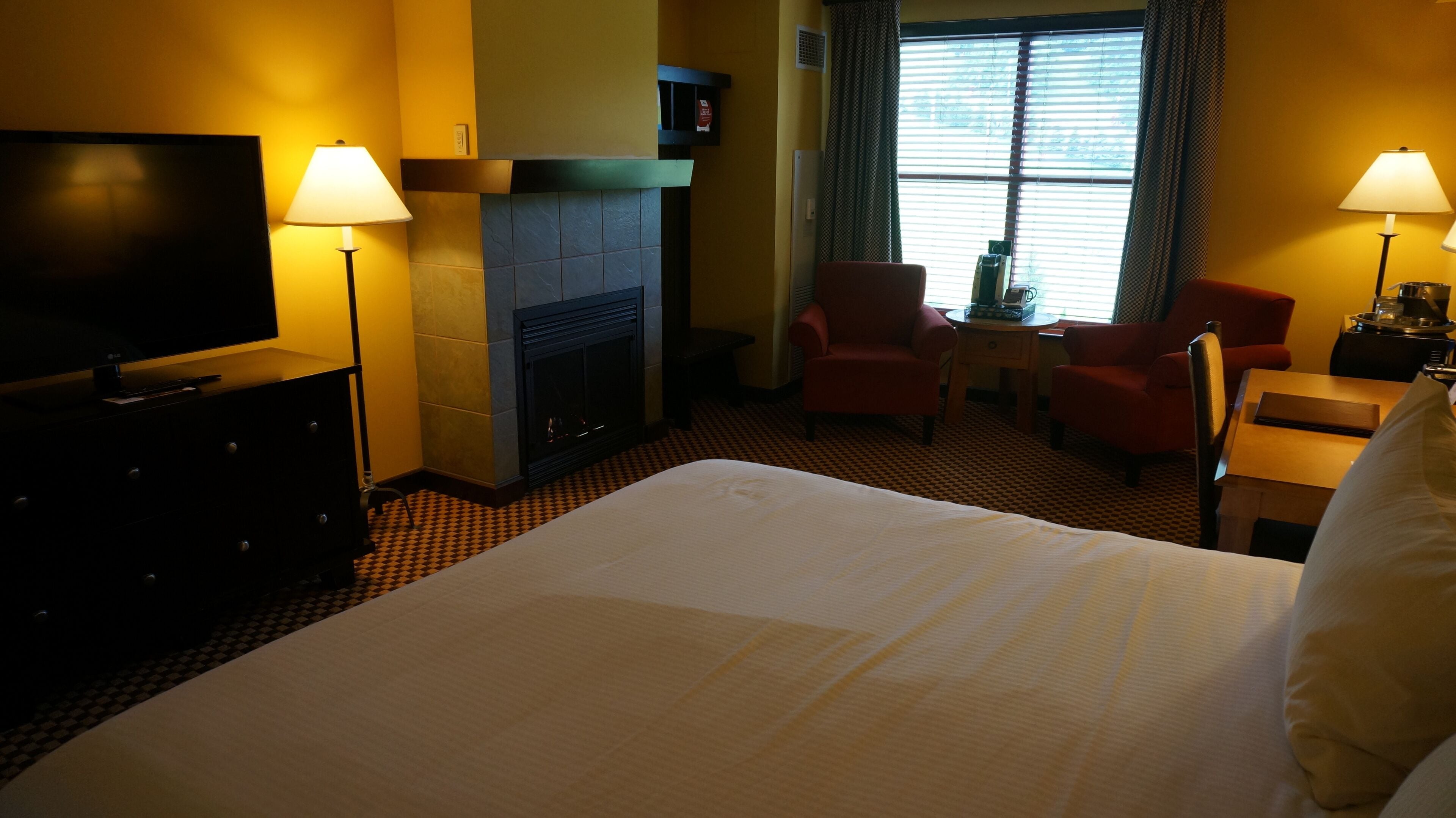 A hotel room with a large bed, a television on a dresser, two chairs, a small table, and a fireplace.