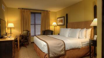 A hotel room with a large bed, two bedside lamps, a desk with a chair, a window with blinds, and a framed picture on the wall.