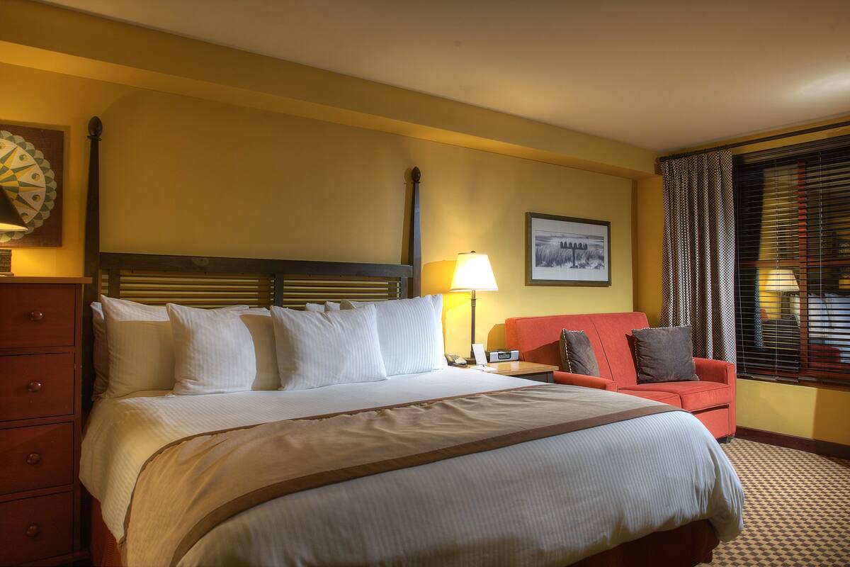 Een hotelkamer met een groot bed, een bureau, een stoel, een dressoir, een lamp en een raam met jaloezieën.