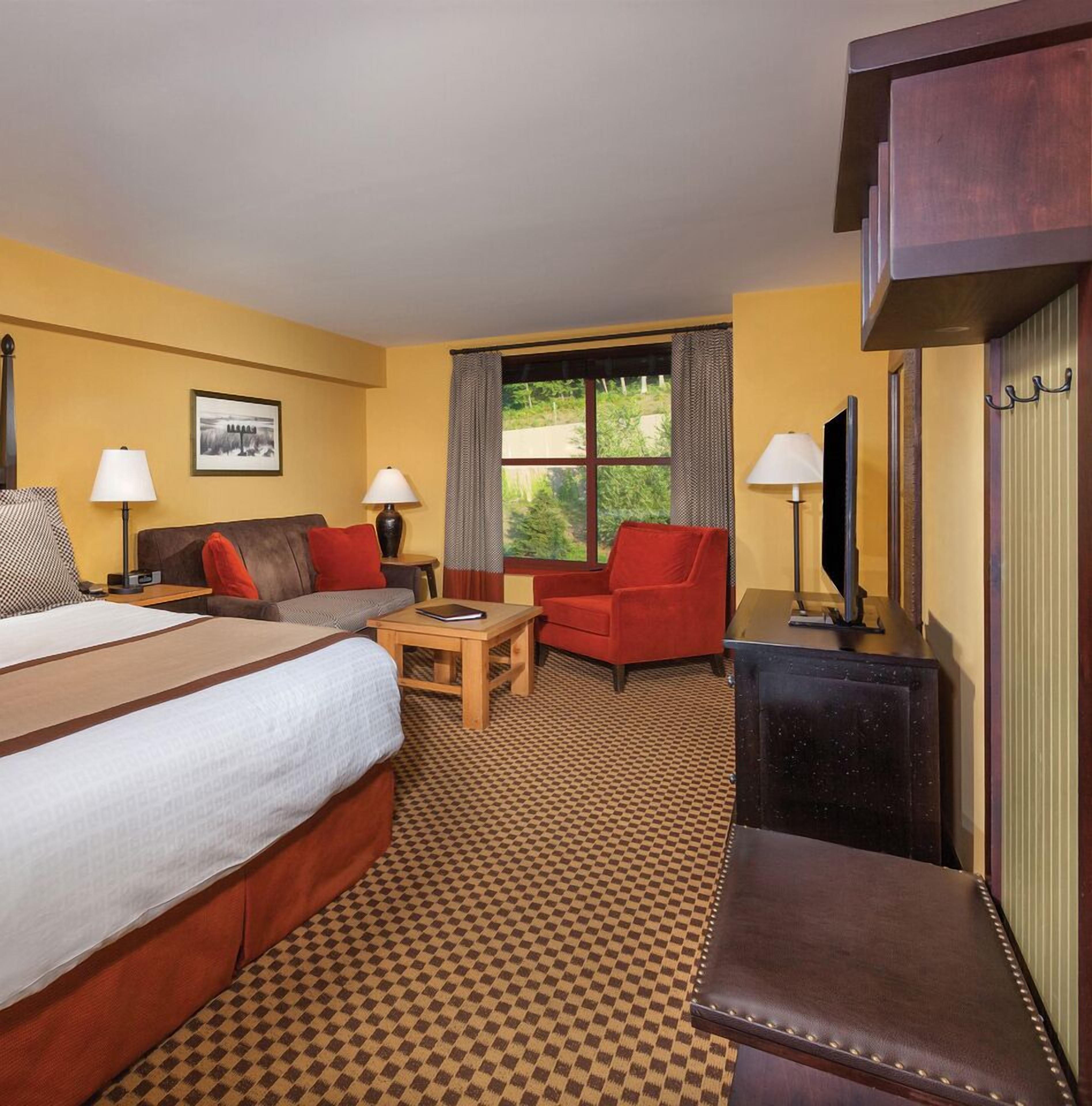 Une chambre d’hôtel avec un grand lit, deux fauteuils, une petite table, un téléviseur et une fenêtre avec des rideaux.