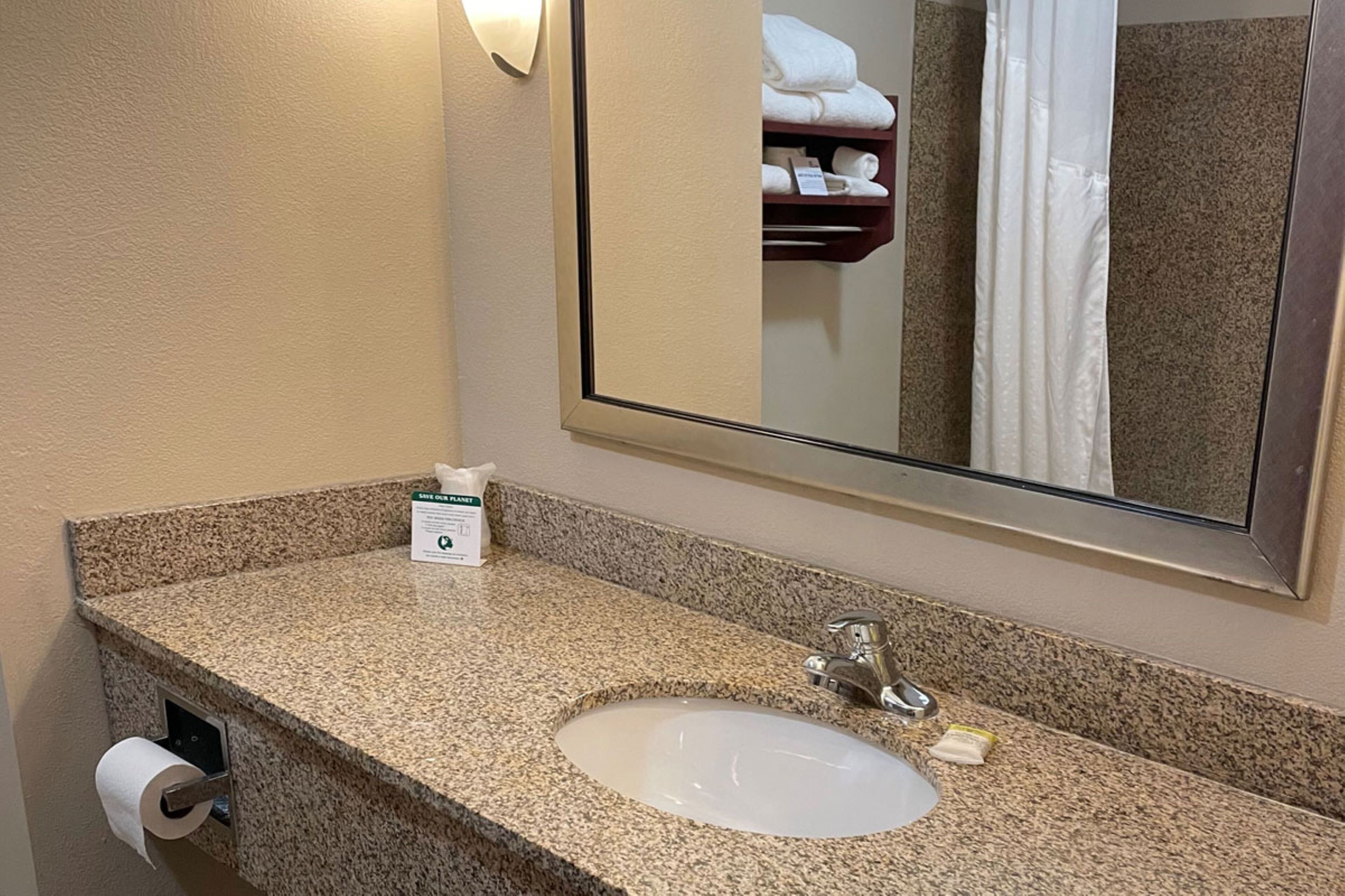 Un baño de hotel con encimera de granito, lavabo, espejo, toallero y dispensador de papel higiénico.
