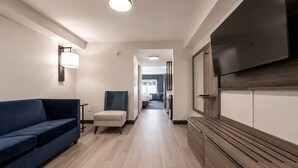 A hotel corridor with a blue sofa, a chair, a TV mounted on a wooden cabinet, and a hallway leading to a bedroom. - Holiday Inn Express Hotel & Suites Niagara Falls by IHG (Niagara Falls)