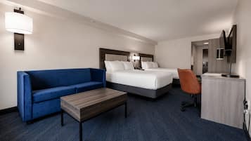 A hotel room with a blue sofa, a wooden coffee table, a bed with white linens, an orange chair, and a desk with a lamp.