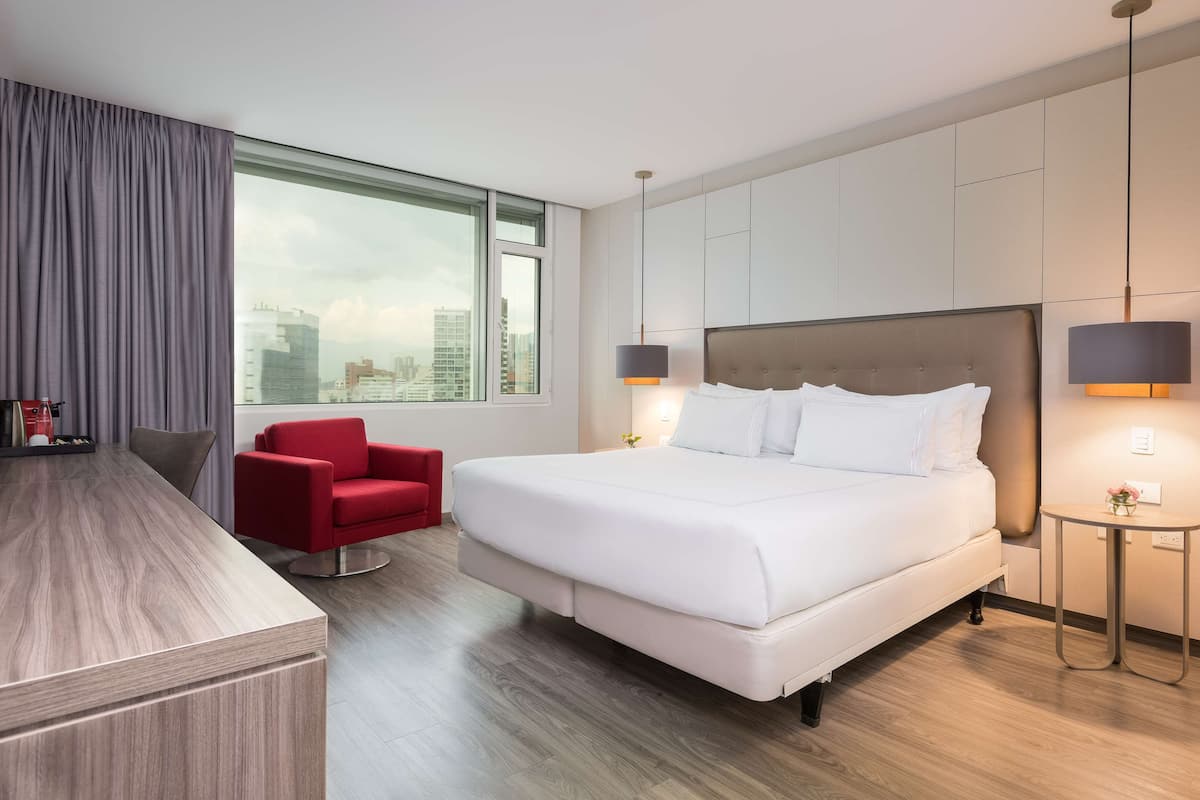 Hotellihuone, jossa on suuri sänky, punainen tuoli, puinen työpöytä ja suuri ikkuna, josta avautuu näkymä kaupungin siluettiin.