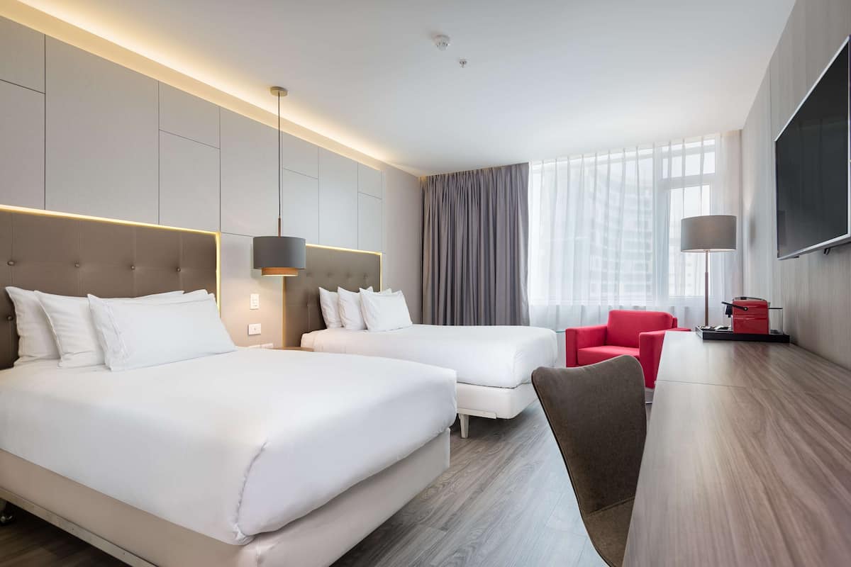Hotellihuone, jossa on kaksi sänkyä, punainen nojatuoli, televisio ja suuri ikkuna, jossa on verhot.