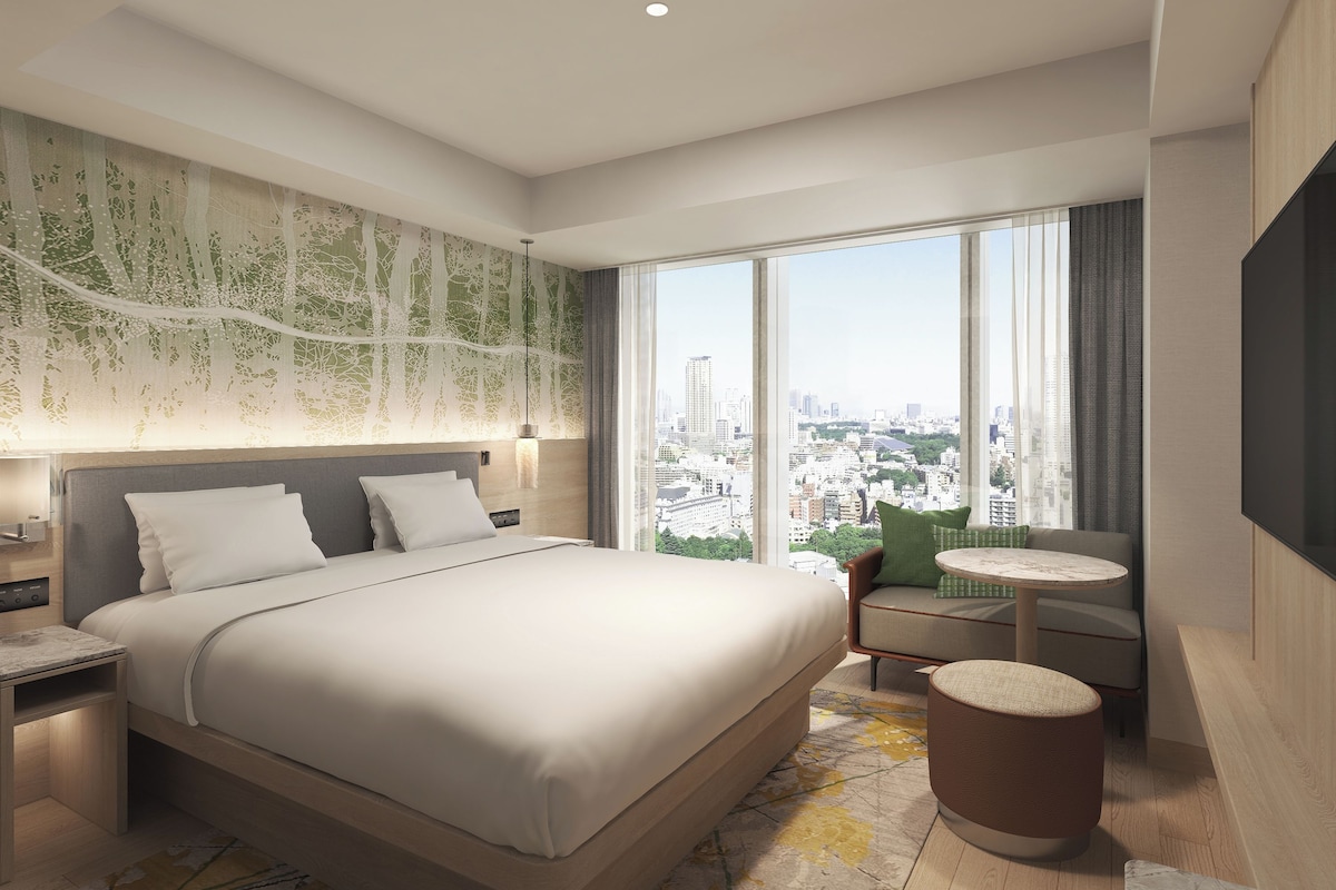 A modern hotel room with a large bed, a city view, a round table, and a TV.