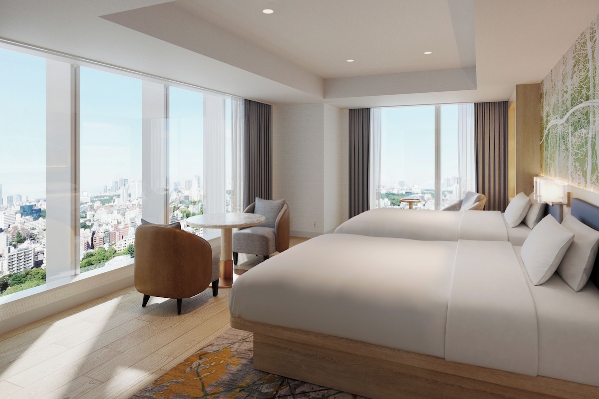 A hotel room with a large bed, a small table, a chair, and a view of the city.