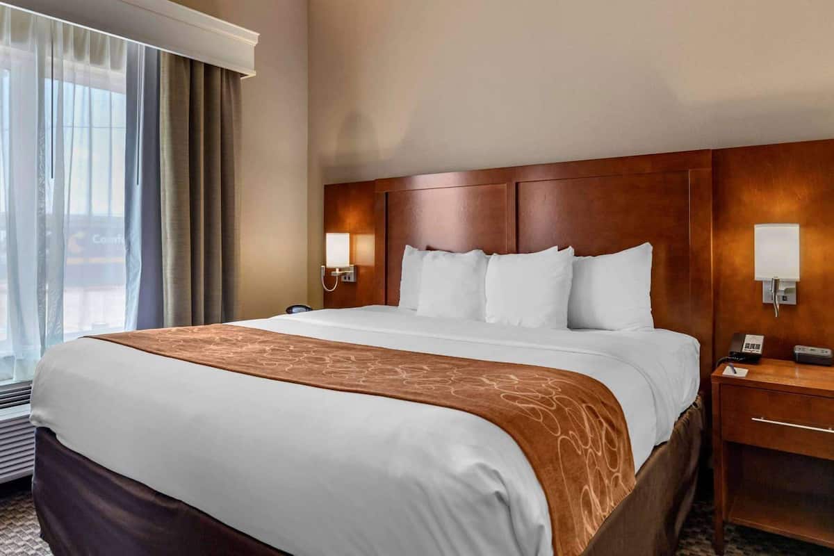 Habitación de hotel con una cama grande, cabecera de madera, lámparas de noche, mesita de noche y una ventana con cortinas.