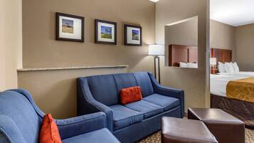 Habitación de hotel con un sofá azul, un sillón más pequeño, un reposapiés marrón y una cama con cabecera. Se observan tres cuadros enmarcados en la pared y una lámpara en el suelo.