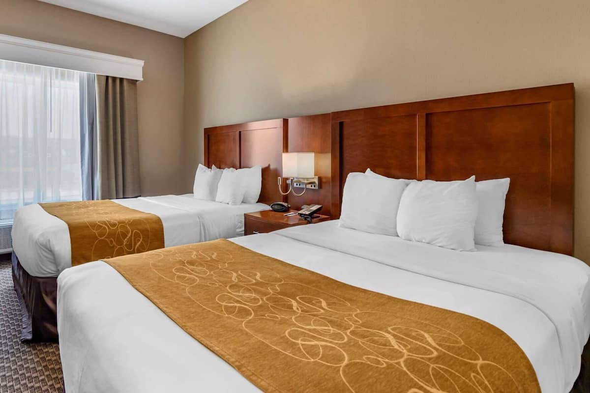 Una habitación de hotel con dos camas, cabeceros de madera, una mesita de noche con lámpara y una ventana con cortinas.