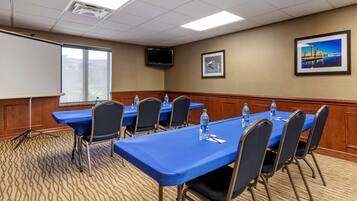 A conference room with a long table, chairs, a projector screen, and a television on the wall.