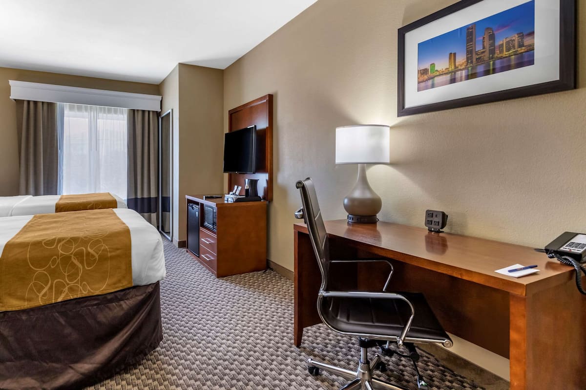 Habitación de hotel con dos camas, un escritorio con lámpara, televisión y un cuadro de paisaje urbano en la pared.