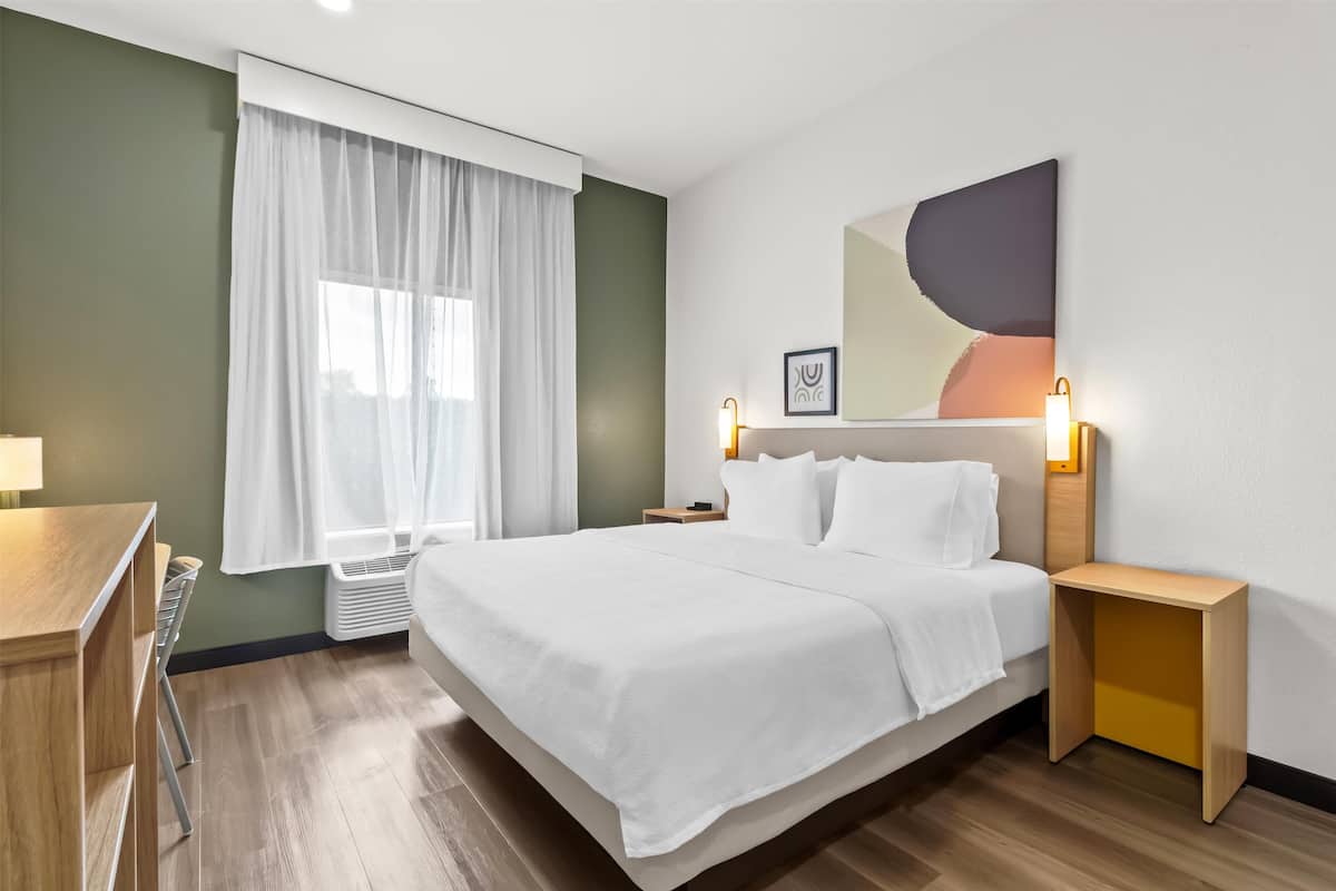 Une chambre d’hôtel avec un grand lit, un bureau, une chaise, une table de chevet, une lampe et une fenêtre avec des rideaux.