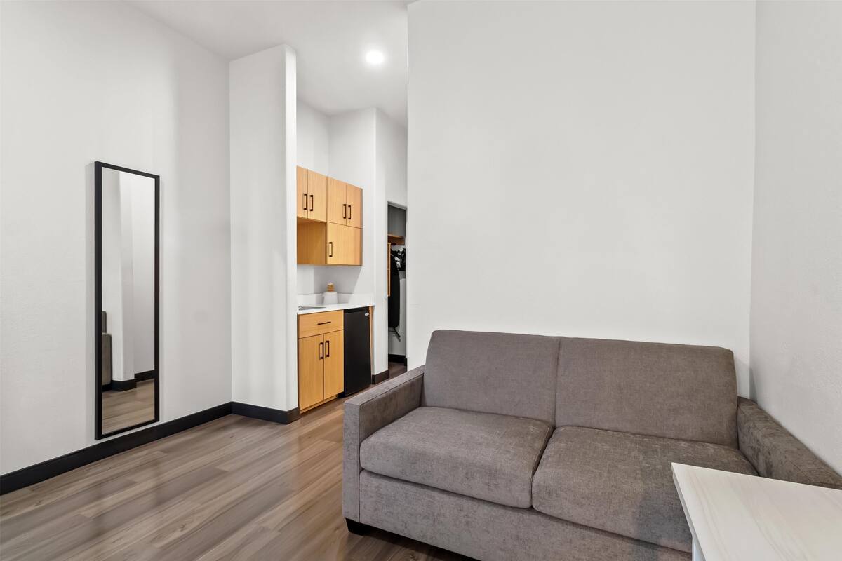 Un salon moderne avec un canapé gris, un plancher en bois et un coin cuisine doté de meubles en bois et d’un réfrigérateur noir.