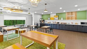 A modern dining area with wooden tables and chairs, a kitchen counter with a 'Delight in the day' sign, and a green wall with artwork.